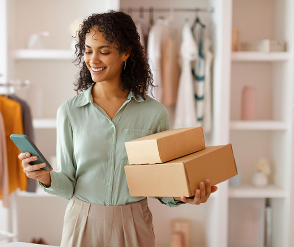 Smiling business owner checking her phone while holding shipping boxes in her retail store